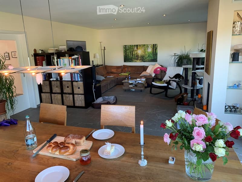 A cozy living and dining area with a brown sectional sofa, coffee table, wooden dining table set for breakfast, and a cat tree. The space features a bookshelf, a rocking chair, and a vase of flowers on the table.