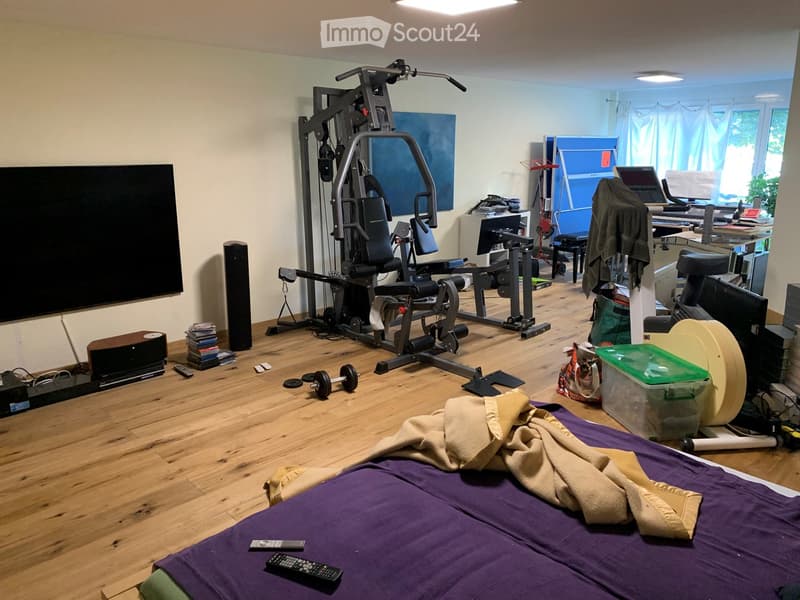 A multi-purpose room with a bed, fitness equipment, a TV, and a desk area with a laptop and other items.