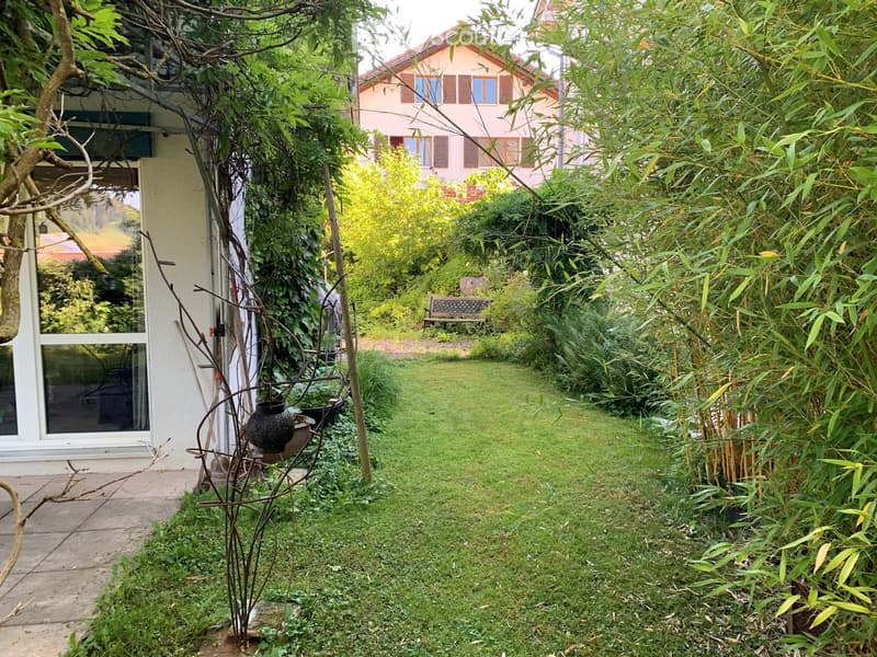 A lush garden with a grassy lawn, various plants, a decorative metal sculpture, a bench, and a pathway leading to a house entrance.