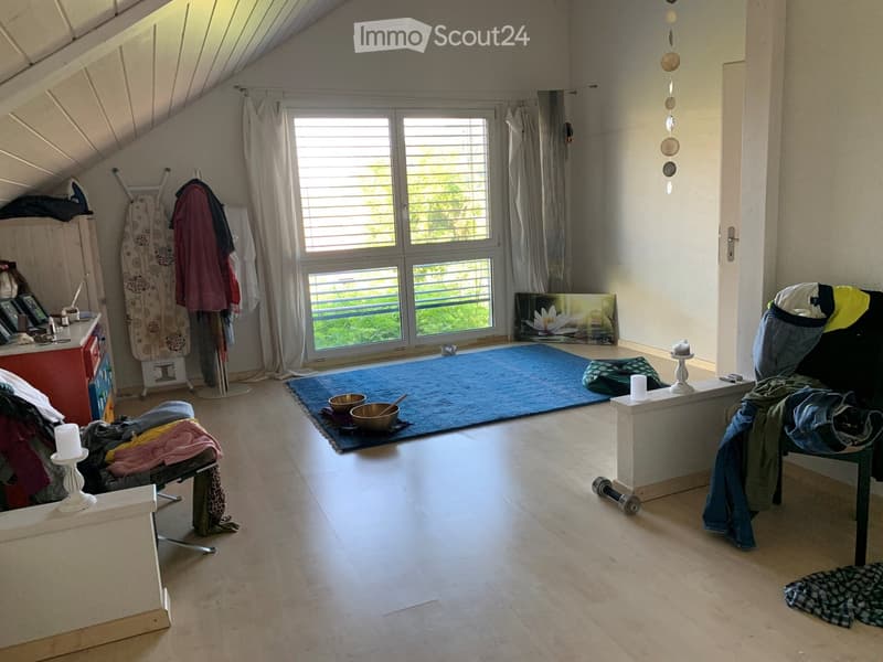 A cozy attic room with wooden floors, large windows, a blue yoga mat, and various items including a chair, table, candles, and a picture frame.