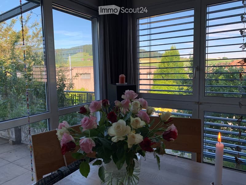 Bright room with large windows offering a scenic view, featuring a wooden table with a vase of roses and a lit candle.