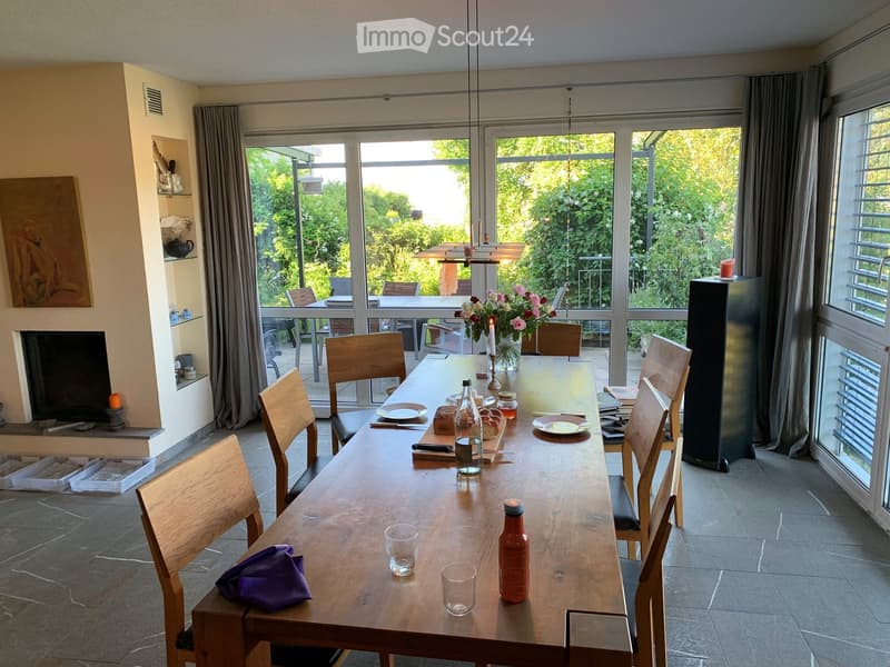 Bright kitchen with large windows offering a view of greenery, a long wooden dining table set with plates, glasses, bottles, and a vase of flowers, surrounded by wooden chairs.