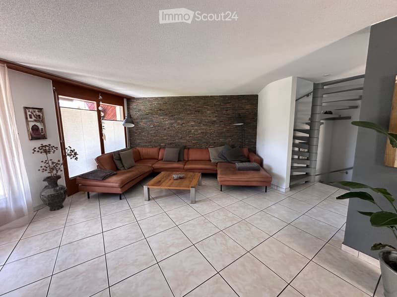 Modern living room with a brown leather sectional sofa, wooden coffee table, decorative vase, and picture frame on the wall. The room features a stone accent wall and a spiral staircase leading to an upper level.