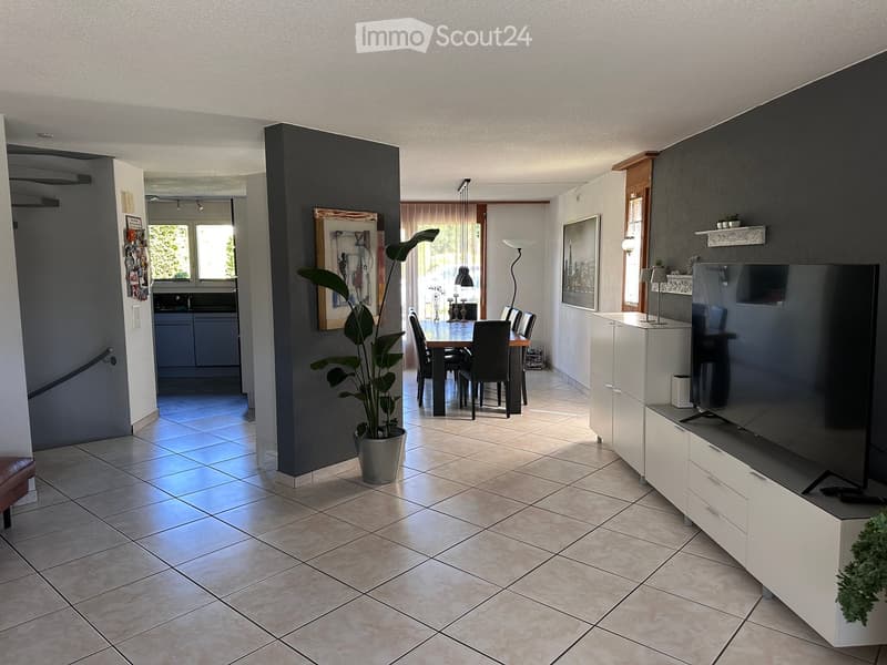 A modern living room with tiled flooring, a large flat-screen TV on a white cabinet, a dining area with a table and chairs, and a potted plant in the corner.