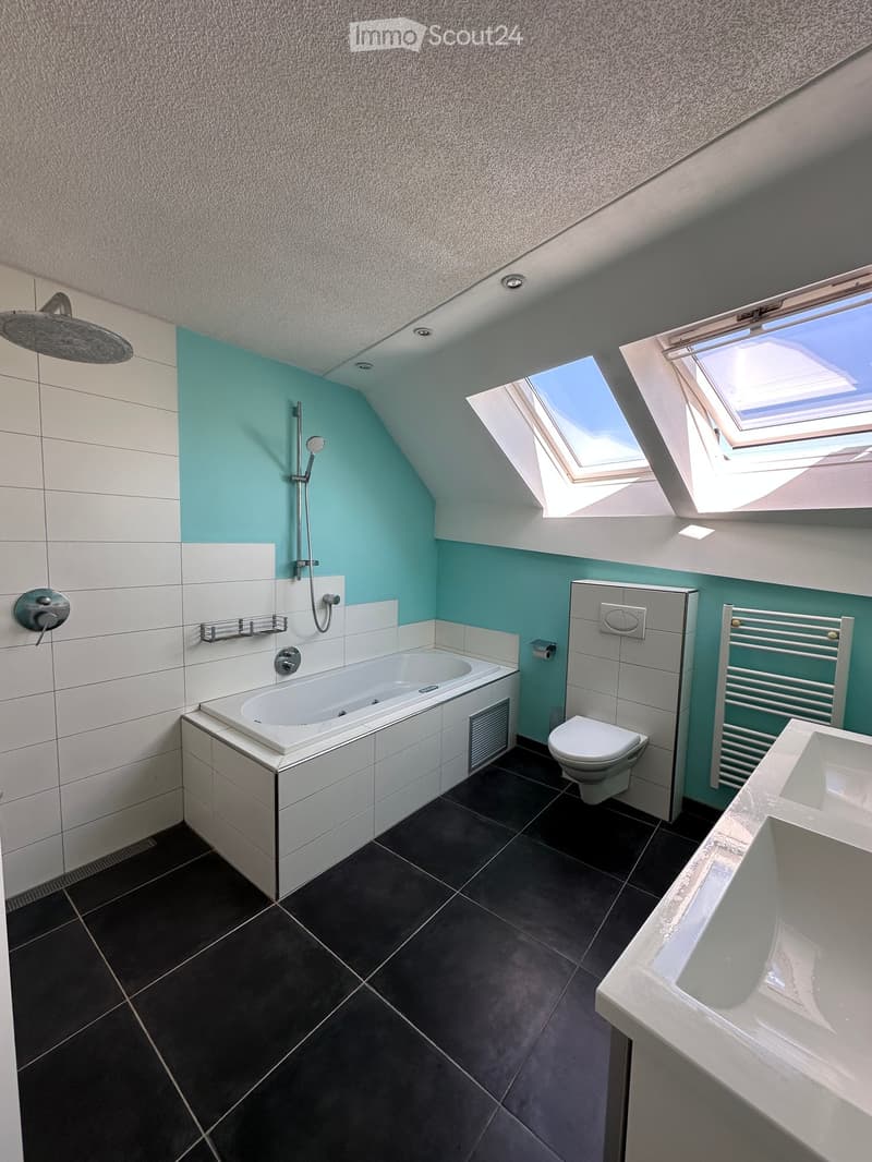 Modern bathroom with a bathtub, toilet, sink, shower, and radiator. The walls are tiled in white and light blue, and the floor is dark tiled. The ceiling has skylights.
