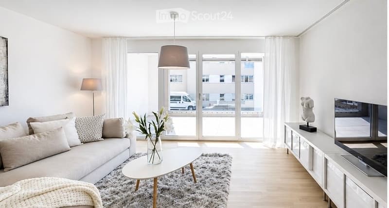 Modern living room with large windows, white walls, grey sofa, coffee table, floor lamp, TV, sideboard, and decorative vase.