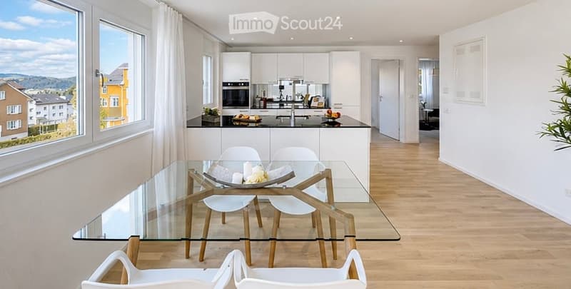 Bright, modern kitchen and dining area with white cabinets, a glass dining table with white chairs, and a kitchen island with an oven and sink. Large windows offer a scenic view of the surrounding area.