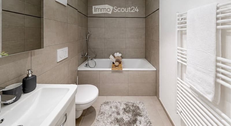 Modern bathroom with a white tiled design, featuring a sink, toilet, bathtub, mirror, towel rack, and a small rug on the floor.