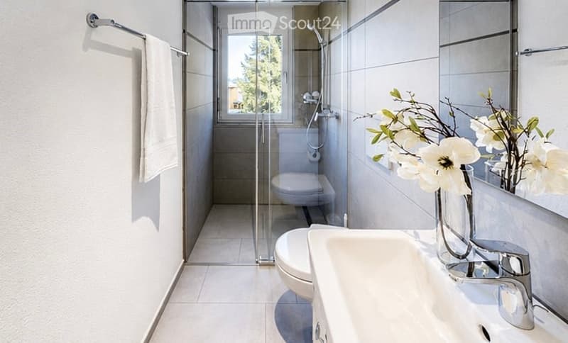 Modern bathroom with white tiles, a glass-enclosed shower, a sink with a chrome faucet, a mirror, a towel on a rack, and a vase with white flowers on the sink counter.