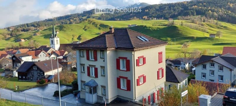 A multi-story residential building with red shutters and a tiled roof, situated in a picturesque village with green hills and a church in the background.