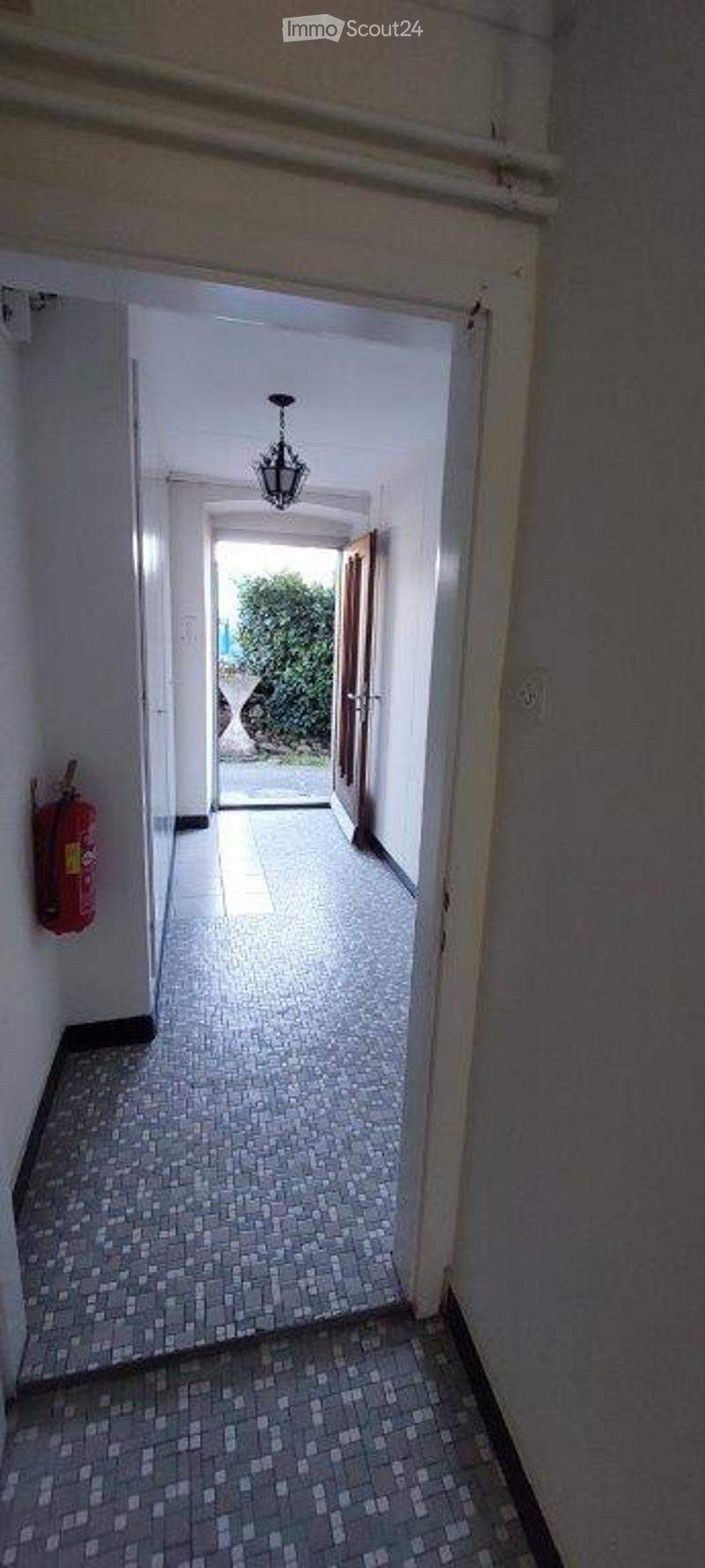 A narrow hallway with tiled flooring, white walls, and a chandelier hanging from the ceiling. A red fire extinguisher is mounted on the left wall, and an open doorway at the end leads to another room.