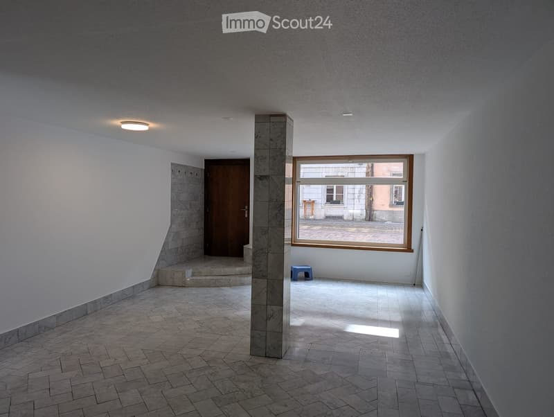 Empty hallway with tiled floor, white walls, a central column, a large window, and a small blue stool near the window.