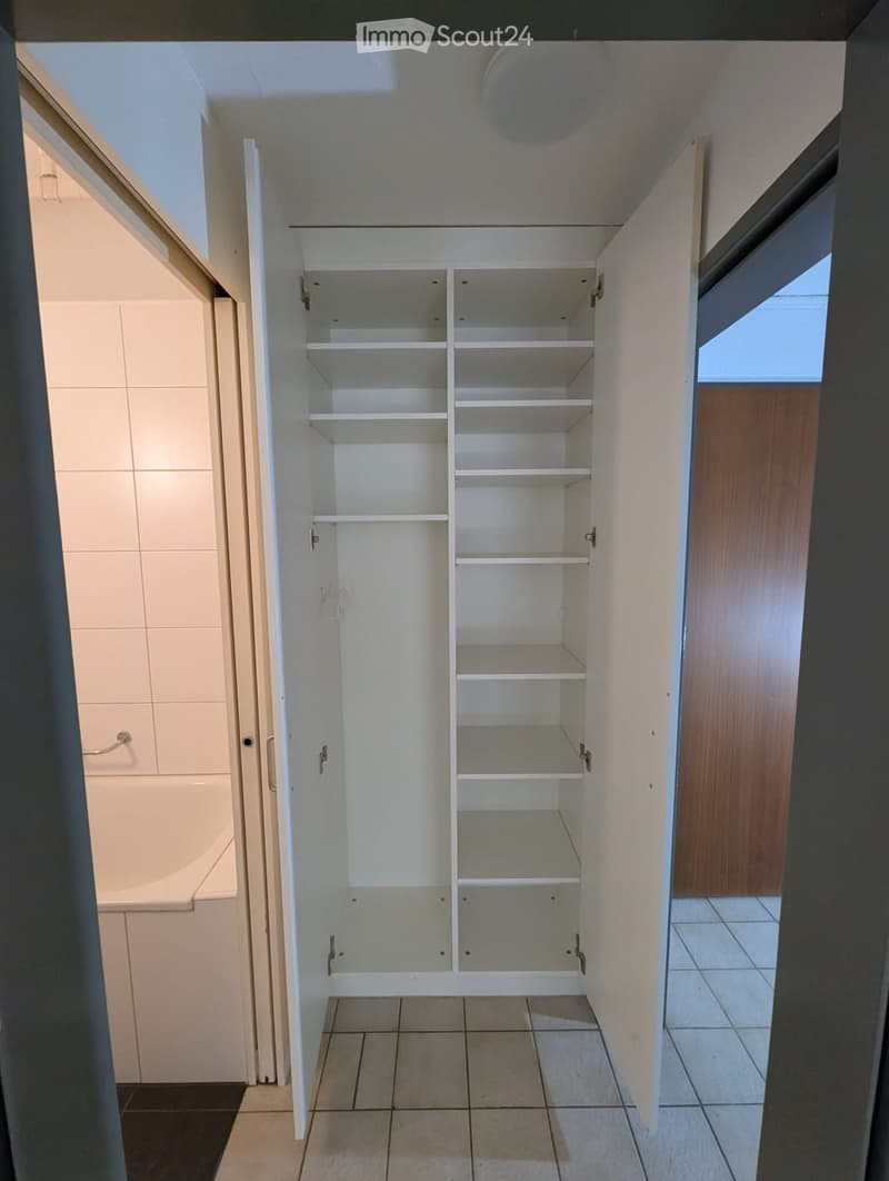 Empty closet with white shelves and beige tiled floor, adjacent to a tiled bathroom area with a bathtub and wooden door.