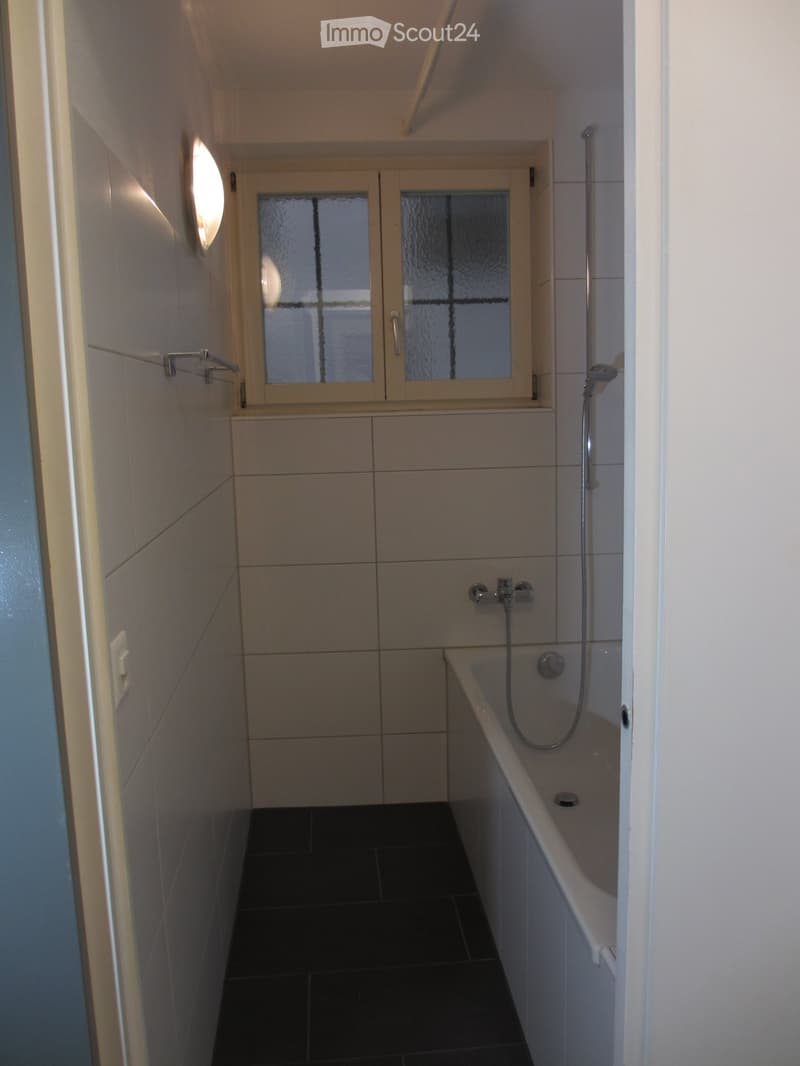 Modern bathroom with a bathtub, shower, and frosted glass window. Features dark tiled flooring and light-colored tiled walls.