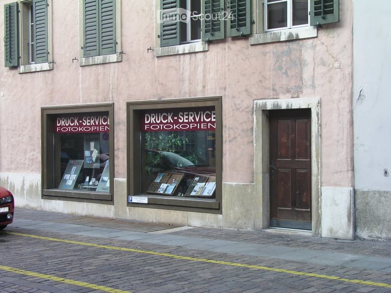Facade of a two-story building with green shutters, large windows, and a storefront displaying signs for 'DRUCK-SERVICE FOTOKOPIEN'.