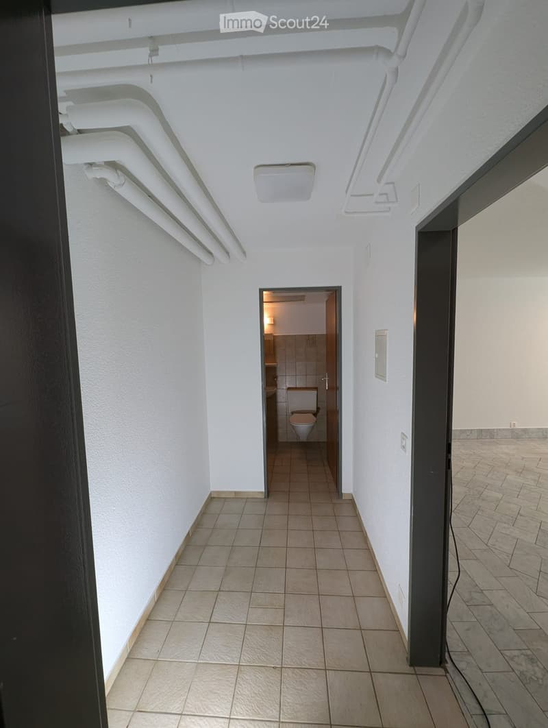 A narrow hallway with white walls and tiled flooring, leading to a small bathroom with a toilet and sink.