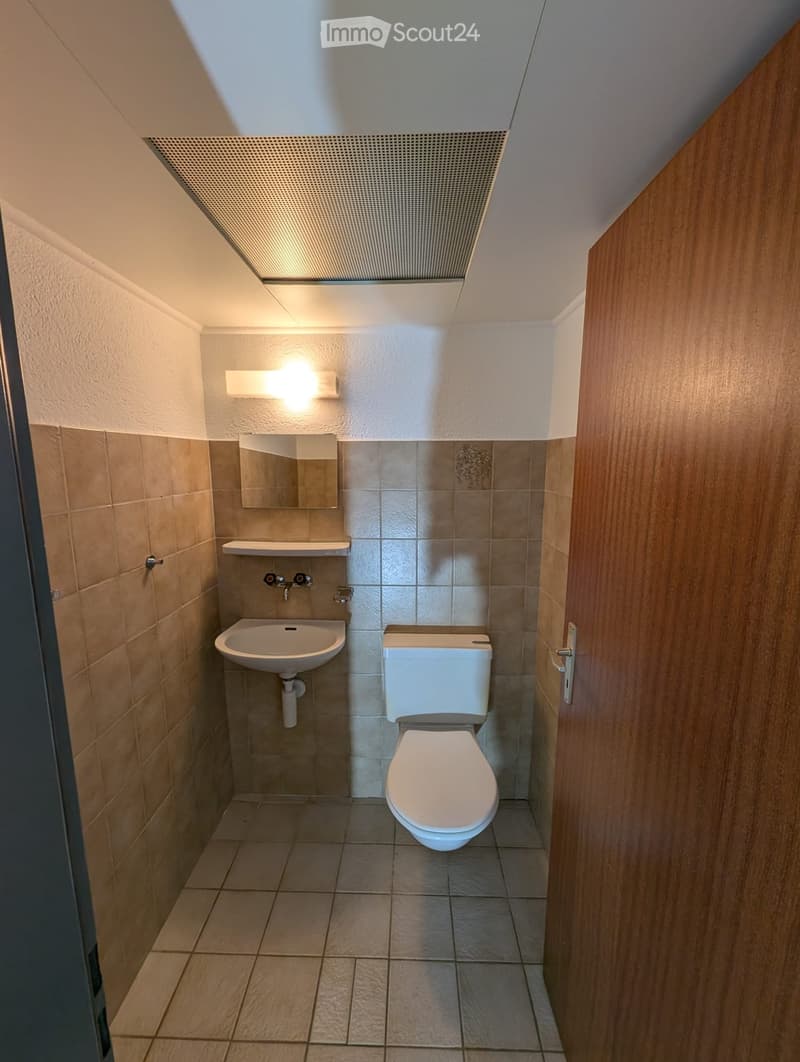 Modern bathroom with beige tiles, white toilet, sink, and mirror. Features a sleek, contemporary design with a built-in shelf above the sink.