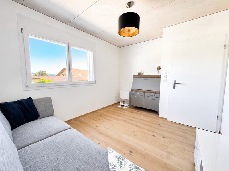 Modern living room with light wooden flooring, white walls, large windows offering a view, grey sofa with dark blue cushions, pendant light, small cabinet, and a robot vacuum on the floor.