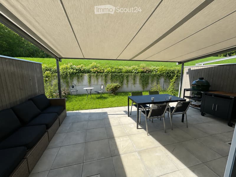 A modern outdoor patio with a tiled floor, a large retractable awning, a wicker sofa, a dining table with chairs, and a grill. The area is surrounded by a low wall with climbing plants and a well-maintained lawn.