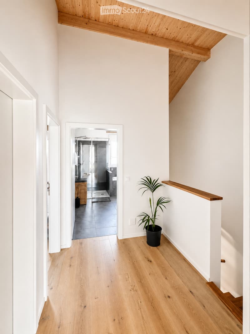 Bright hallway with wooden flooring, white walls, and a wooden railing leading to a staircase. A potted plant is placed near the railing, and an open doorway reveals a modern bathroom with dark tiles and a glass shower enclosure.