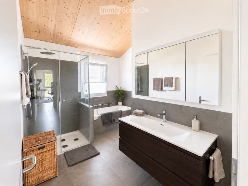 Modern bathroom with a large mirror, sink, bathtub, and glass-enclosed shower. Features include a wicker basket, towels, and a sleek wooden vanity.