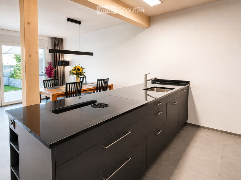 Modern kitchen with dark grey cabinets, a central island, and a dining area with a wooden table and chairs. The kitchen features a sleek sink and faucet, and there are vases with flowers on the table.