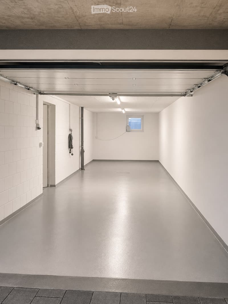 Empty garage with white brick walls, concrete floor, and a large overhead door. Features a small window and a single electrical outlet.