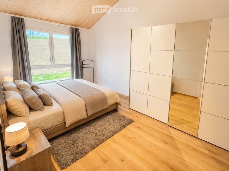 Cozy bedroom with a large bed, nightstand with lamp, and a modern wardrobe. The room features wooden flooring and large windows with curtains, offering a view of a green landscape.