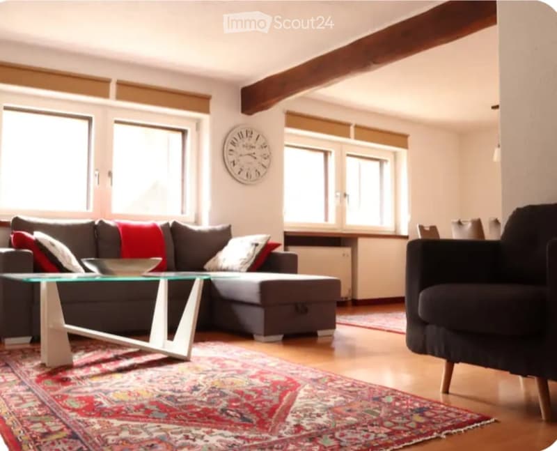 Modern living room with large windows, exposed wooden beams, grey sectional sofa, glass coffee table, armchair, and decorative rug.