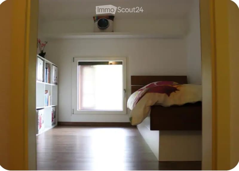 Cozy bedroom with a wooden bed, colorful bedding, a bookshelf, and a window letting in natural light.