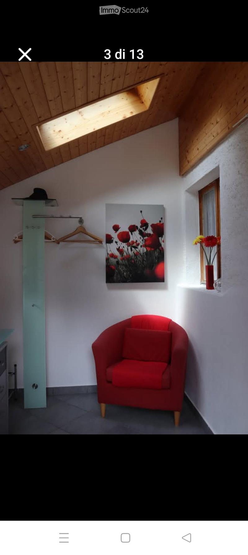 A small, well-lit room with a red armchair, a picture frame on the wall, a vase with flowers on a shelf, and a wardrobe with a coat hanger.