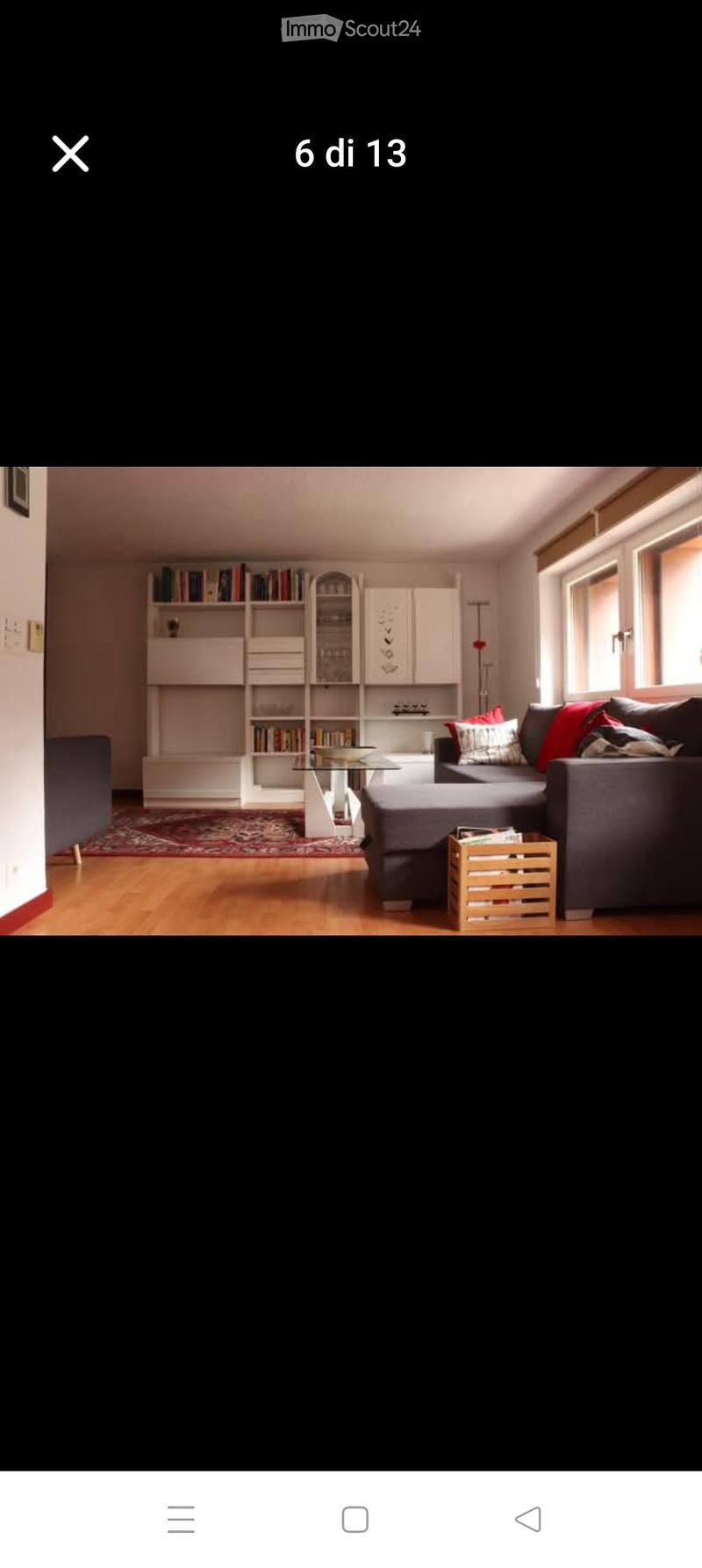 Modern living room with a grey sectional sofa, glass coffee table, bookshelf, and wooden crate. The room features wooden flooring and large windows.