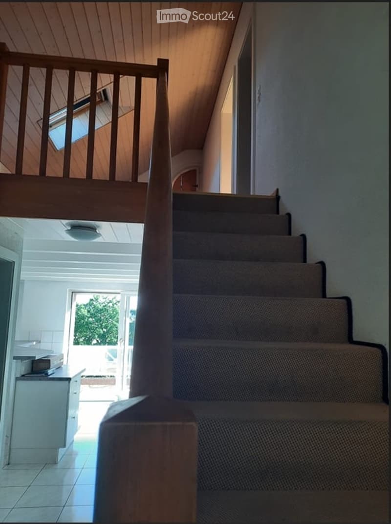 Staircase with wooden handrail and carpeted steps, leading to an upper level with a skylight. The lower level features a kitchen area with white cabinets and a tiled floor.