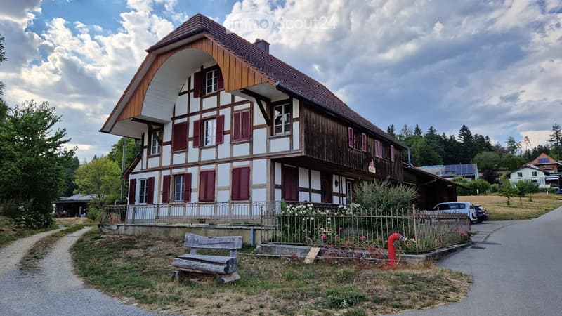 Two-story traditional house with white and brown timber framing, red shutters, and a wooden balcony. The house features a pitched roof and is surrounded by a small garden with a bench and a fence.