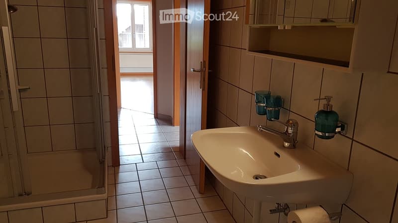 Bathroom with a white sink, chrome faucet, soap dispenser, and a glass shelf holding cups. The room features tiled walls and floor, with a shower area visible through an open doorway.