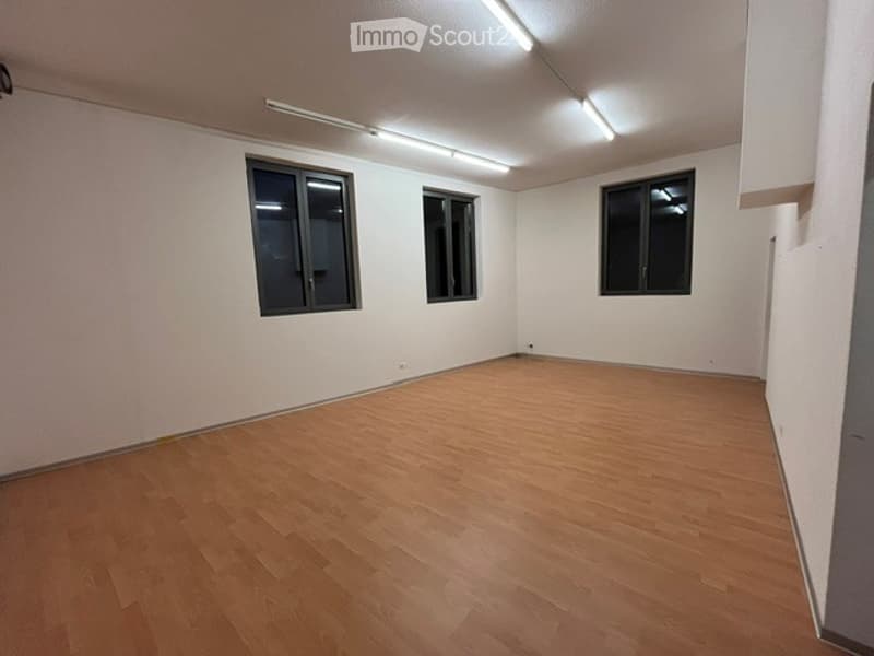 Completely empty room with bare white walls and wooden floor. Three windows are visible, and the room is illuminated by ceiling lights.