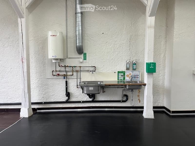 The room has two stainless steel sinks with faucets and a white wall mounted water heater. There are two soap dispensers on the wall next to the sinks and a green box. The pipes are visible on the wall. The room is empty with white walls and black floor.