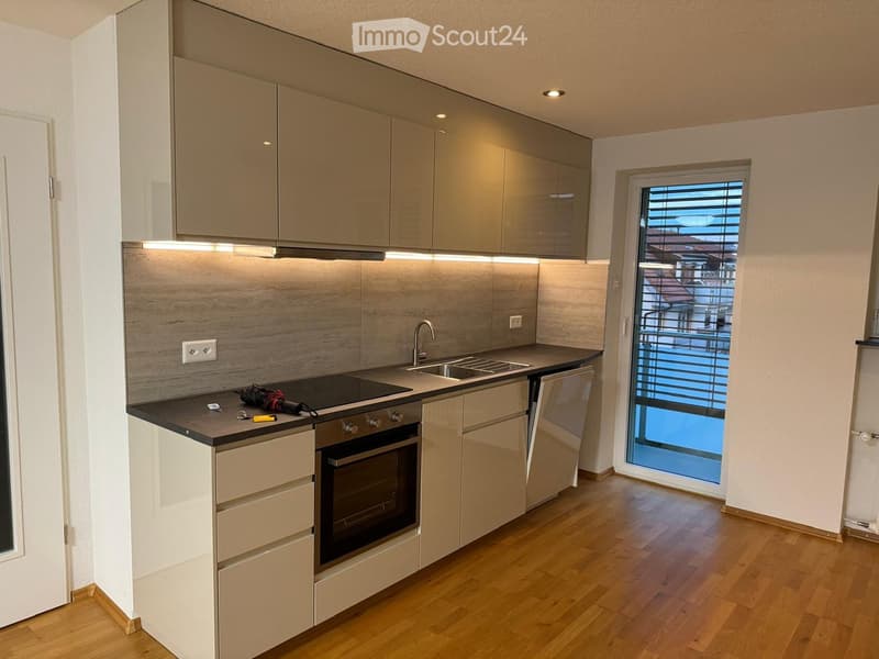 Modern kitchen with light-colored cabinets, a dark countertop, and a built-in oven. Features a sink with a modern faucet, a window with blinds, and a wooden floor.