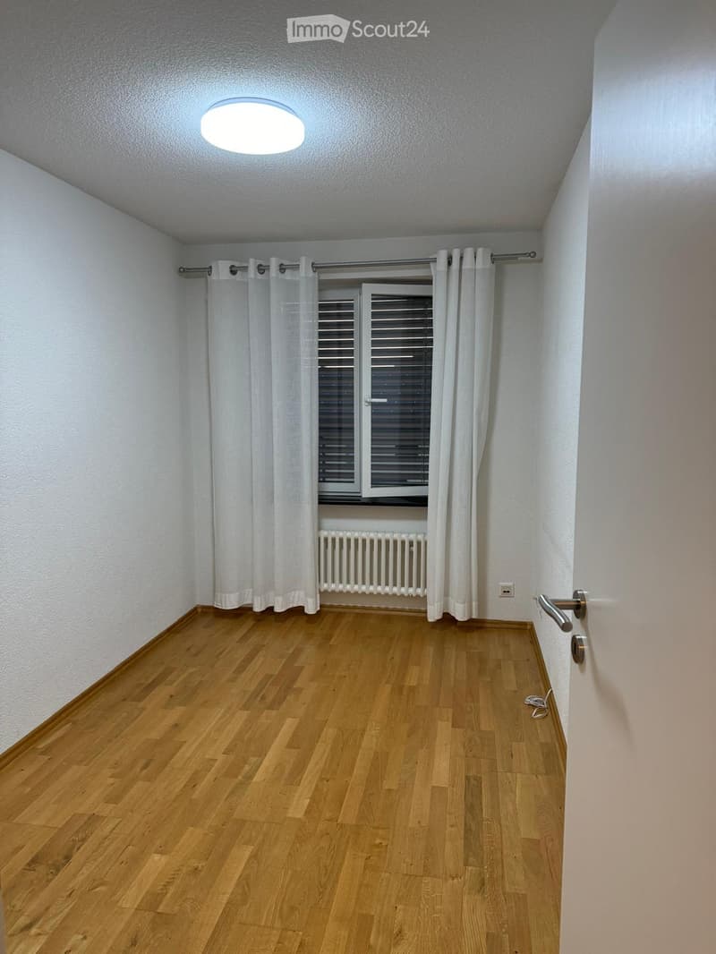 Empty room with wooden flooring, white walls, a window with white curtains, a radiator, and a door with a silver handle.