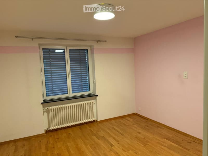 Empty room with wooden flooring, white walls, pink accent wall, window with blinds, radiator, and ceiling light fixture.