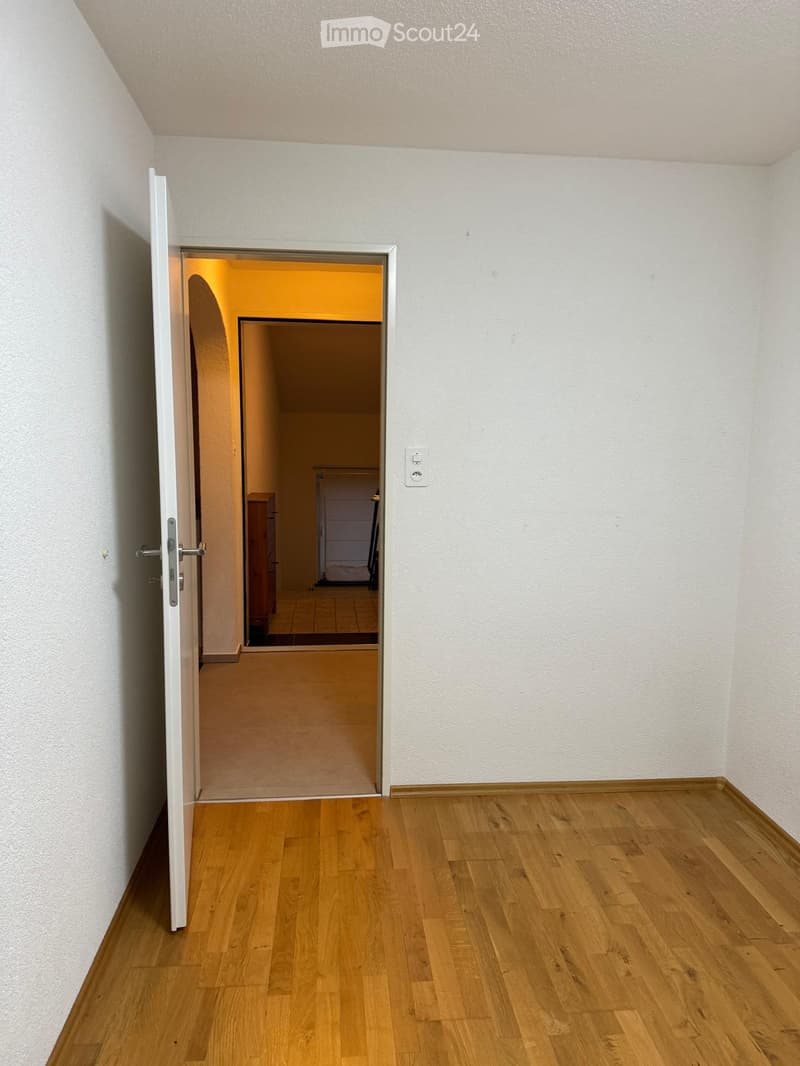 Empty room with wooden flooring and white walls, featuring an open door leading to another room with a visible wardrobe and tiled floor.