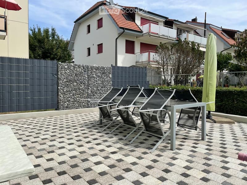 Outdoor patio with checkered tile flooring, a small table, and several chairs arranged for seating. The area is enclosed by a stone wall and a metal fence, with a modern house and balcony visible in the background.