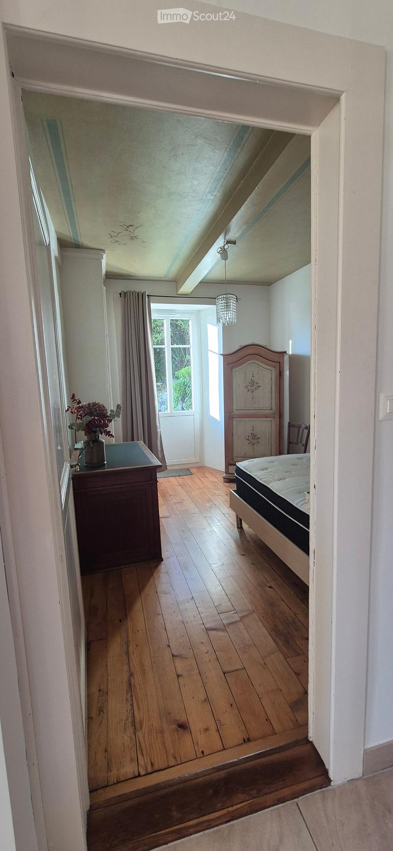 Cozy attic bedroom with wooden floors, slanted ceilings, and large windows. Features a bed, nightstand with a flower vase, a wooden wardrobe, and a chair.