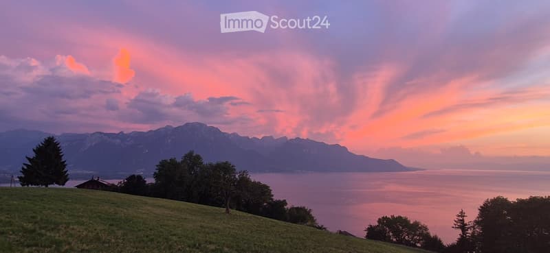 A scenic view of a lake and mountains at sunset, with a small house visible on the left side of the frame.