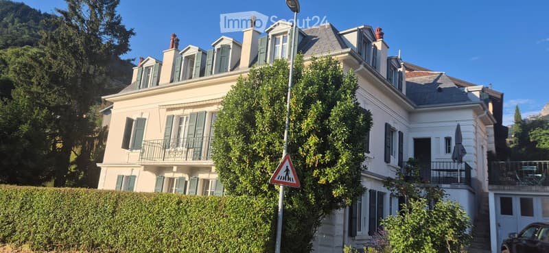 A large, elegant house with multiple stories, white walls, green shutters, and a grey roof. The house features balconies and is surrounded by lush greenery and trees.