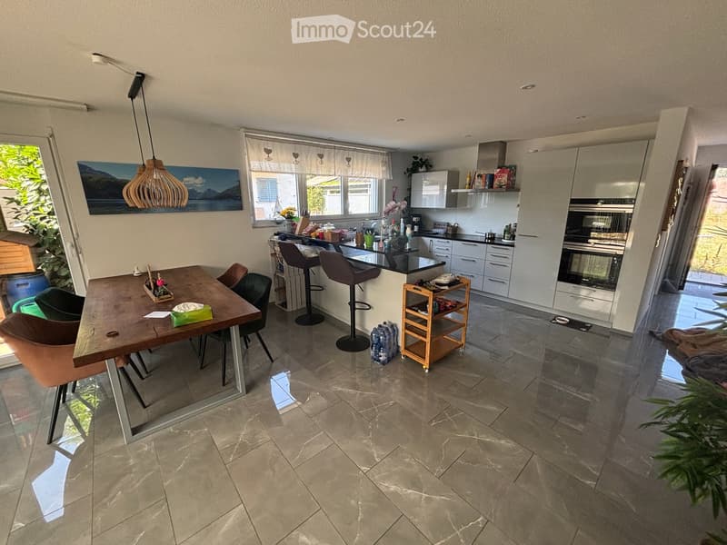 Modern kitchen and dining area with a wooden dining table, chairs, bar stools, kitchen island, built-in oven, refrigerator, and a small kitchen cart. The space features tiled flooring and large windows with curtains.