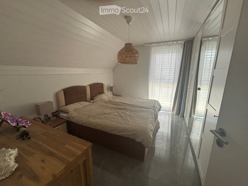 Cozy bedroom with a large bed, nightstand, wooden table, and a hanging woven lamp. The room features white walls, a slanted ceiling, and large windows with curtains.