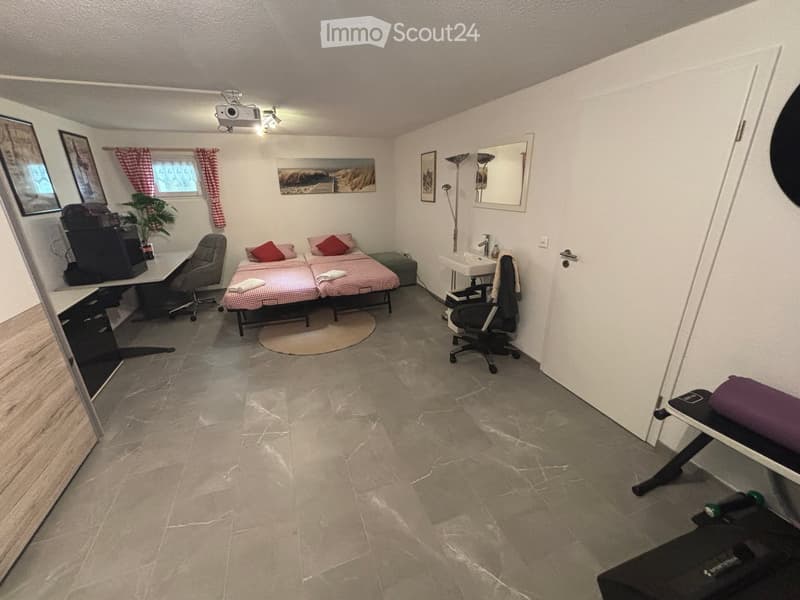A furnished room with a bed, desk, chair, mirror, sink, and plant. The room features a tiled floor and a window with red checkered curtains.