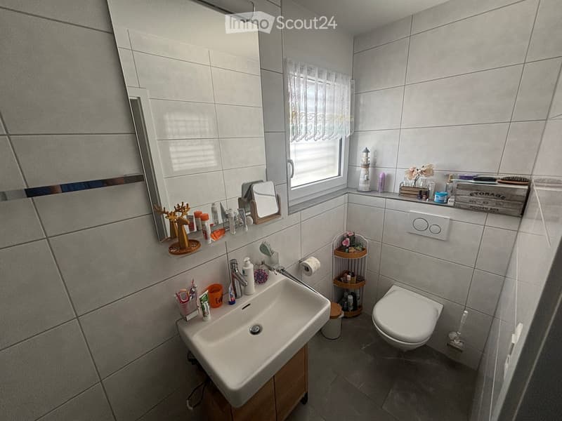 Modern bathroom with white tiles, a sink, toilet, mirror, and various toiletries neatly arranged on shelves and counters.
