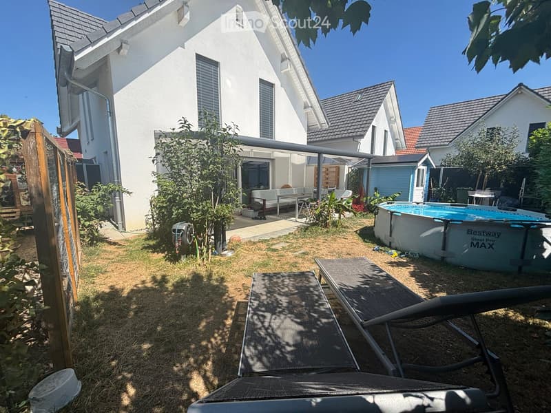 Garden area with sun loungers, a swimming pool, a covered patio with outdoor furniture, and a modern house in the background.